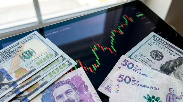 Un primer plano de billetes de cien dólares estadounidenses y billetes de cincuenta mil pesos colombianos sobre una tableta que muestra un gráfico financiero con velas verdes y roj